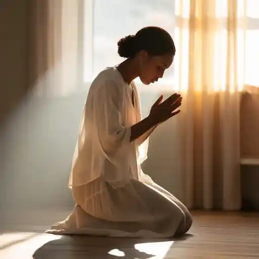 Woman kneeling in prayer beside a bed in a softly lit room with sunlight streaming through a window