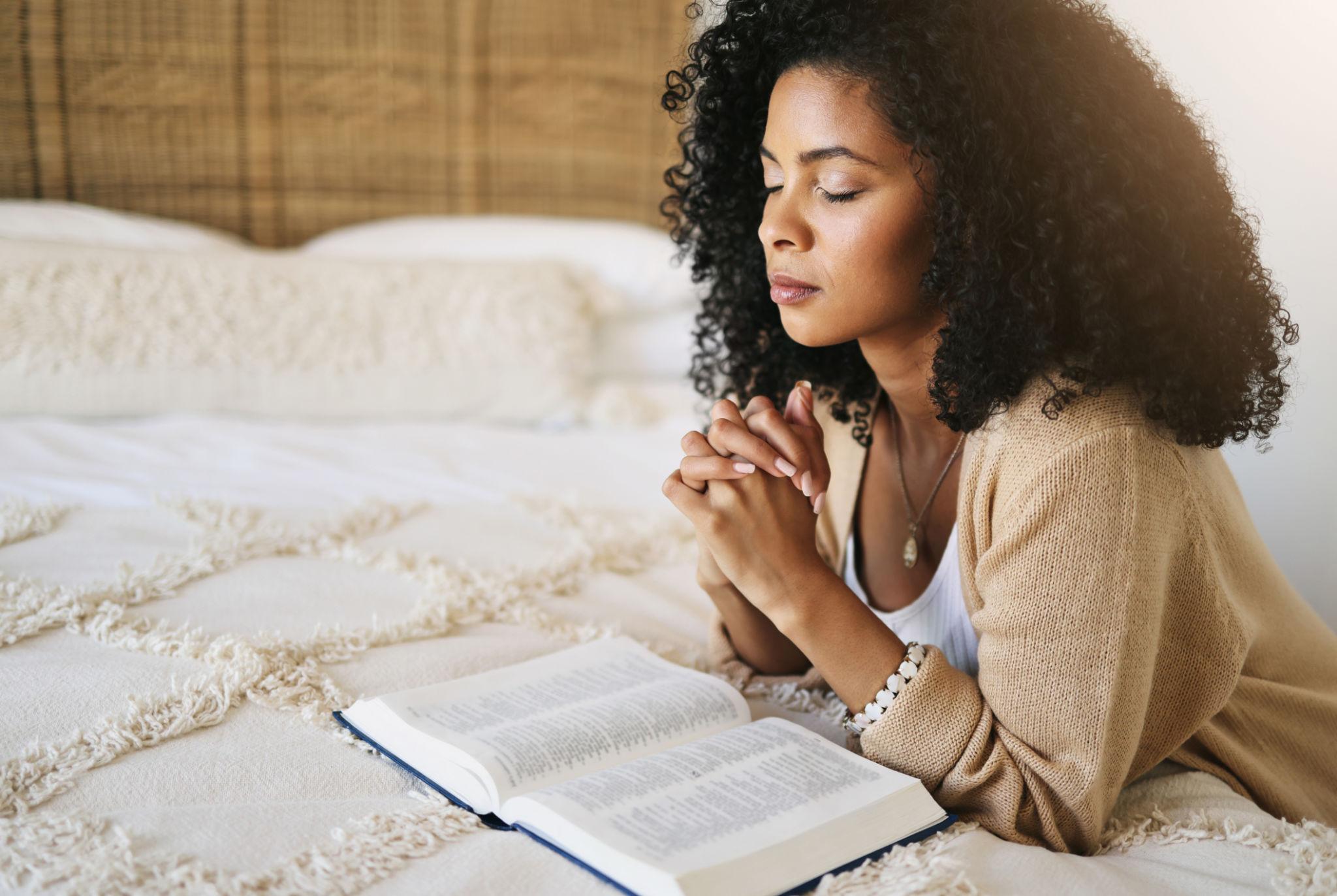 A woman with curly hair lies on a white bed with her eyes closed and hands clasped in prayer over an open Bible.