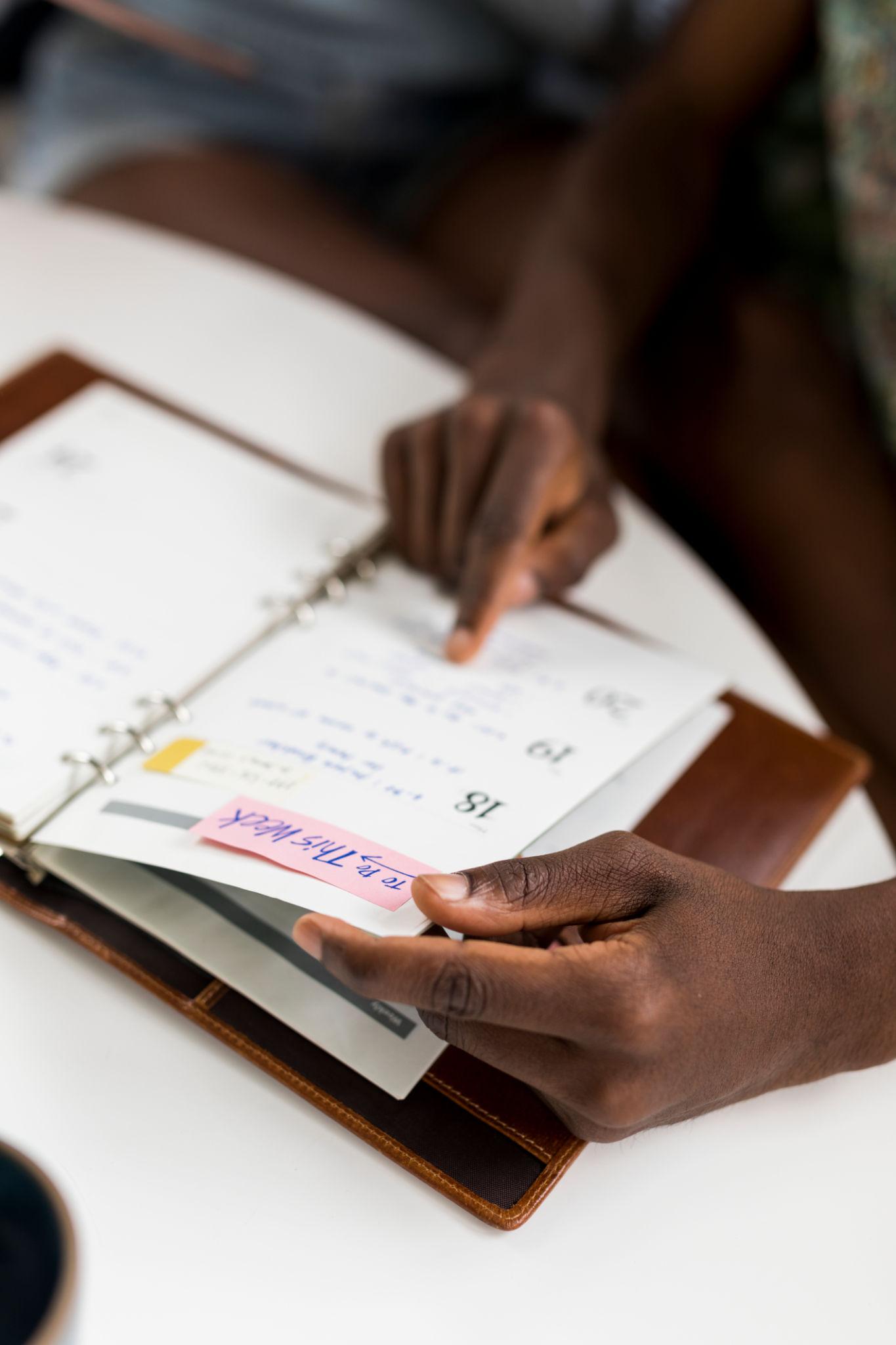 Close-up of a woman's hands pointing to and holding pages in an open leather-bound planner with handwritten notes and a pink sticky note reading "To Do This Week."