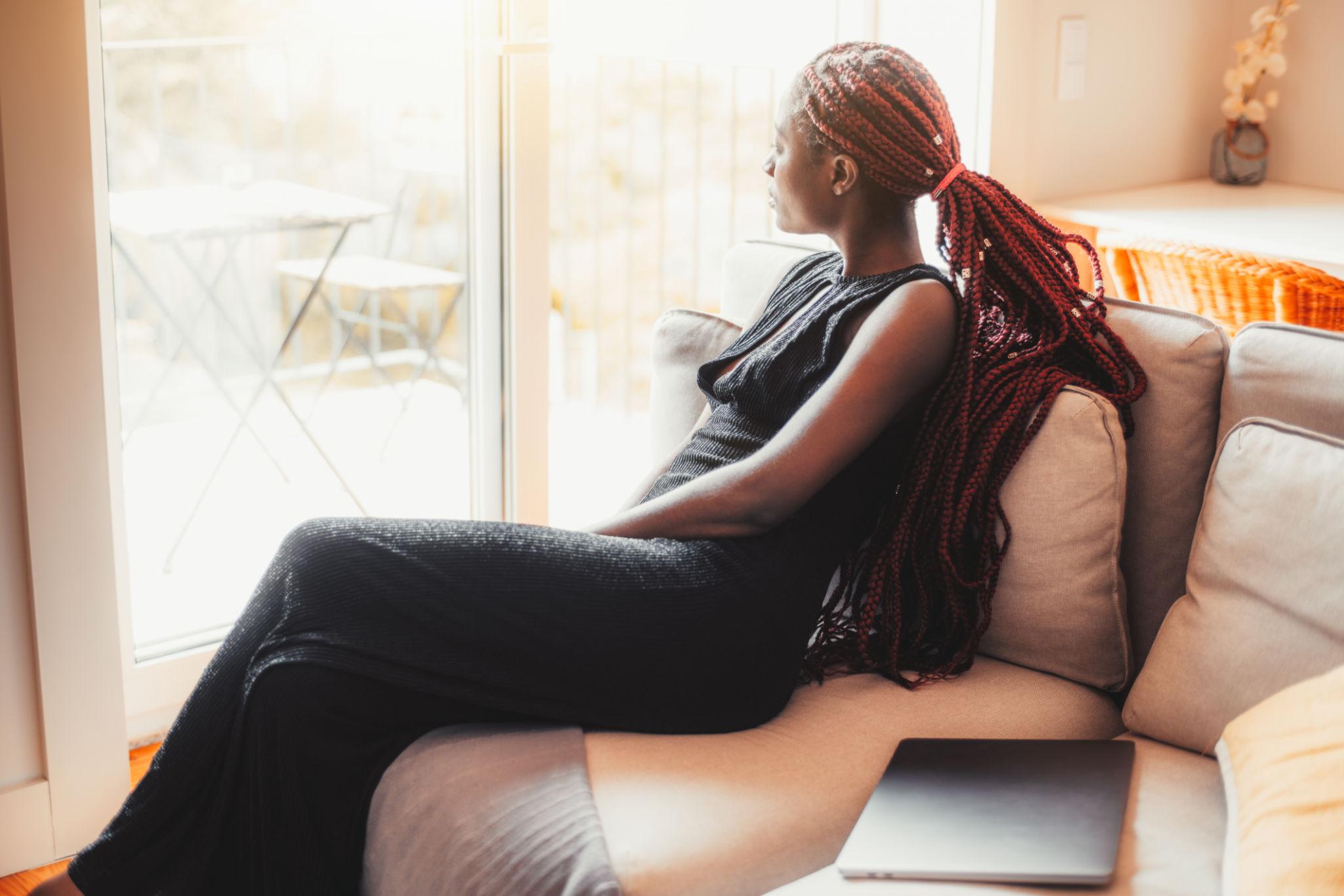 A woman with long braided hair sits quietly on a couch gazing out a sunlit window, a closed laptop beside her — reflecting the stillness of a woman pausing to process her season.