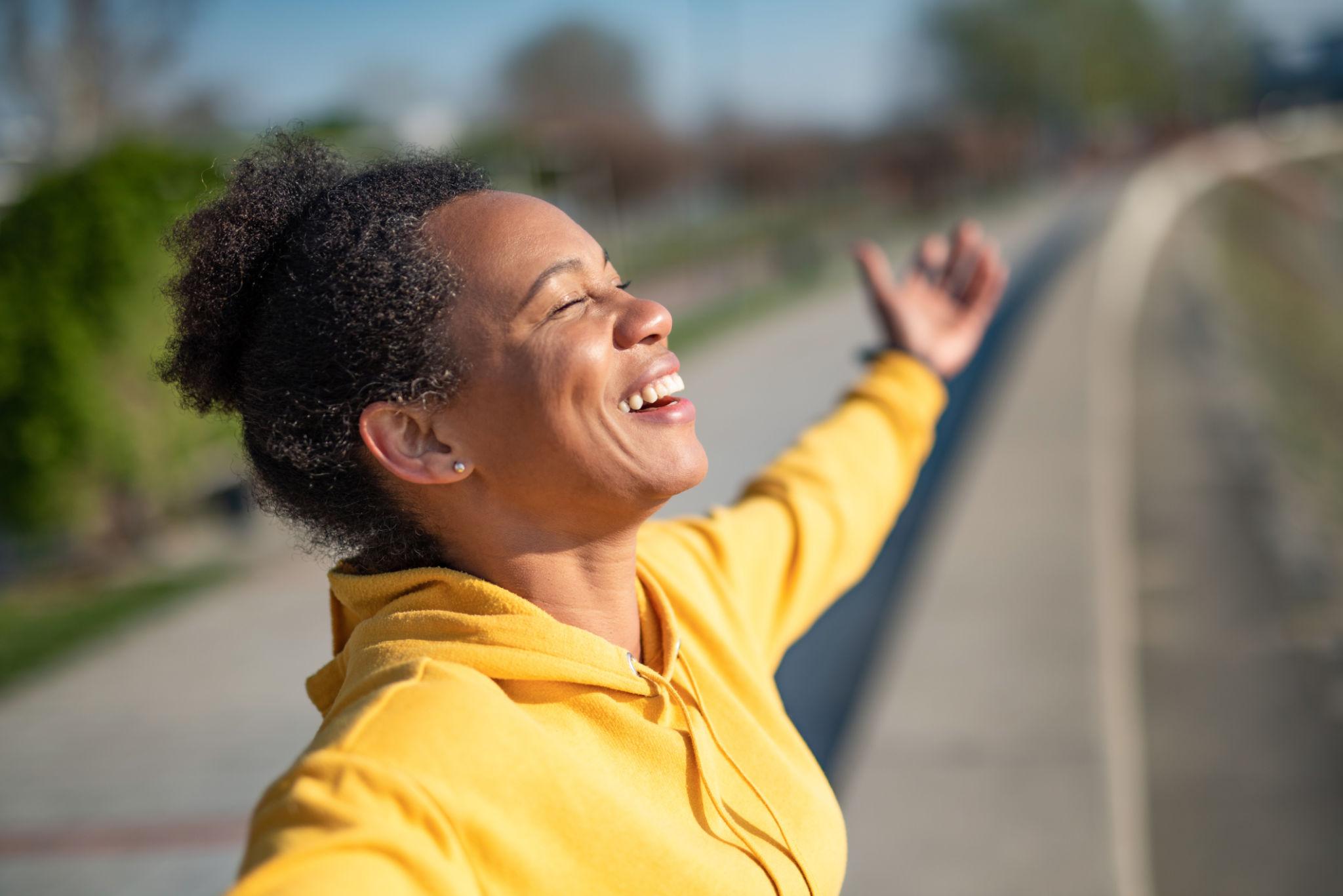 A woman in a yellow hoodie stands outdoors on a path, eyes closed, head lifted, and one arm raised with an expression of pure joy and release.