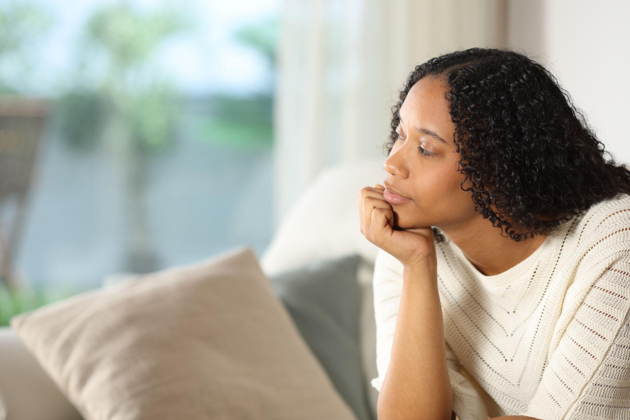 A woman sitting quietly on a couch, chin resting on her hand, gazing thoughtfully into the distance.