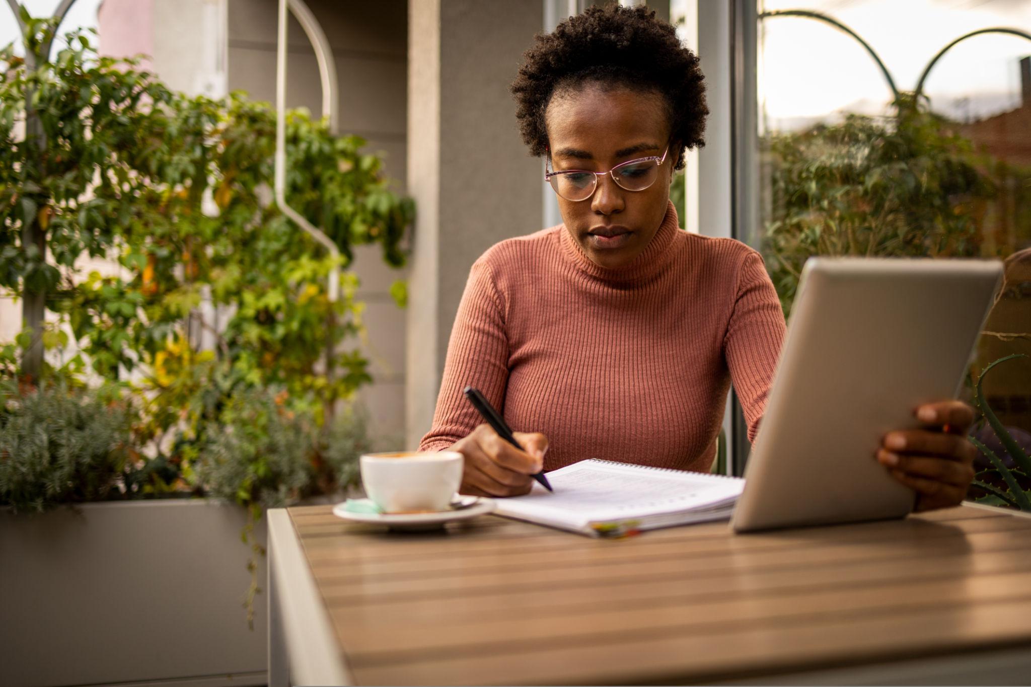 A woman wearing glasses and a rust-colored turtleneck sweater sits at an outdoor table writing in a notebook with a tablet and coffee cup nearby, surrounded by lush green plants.