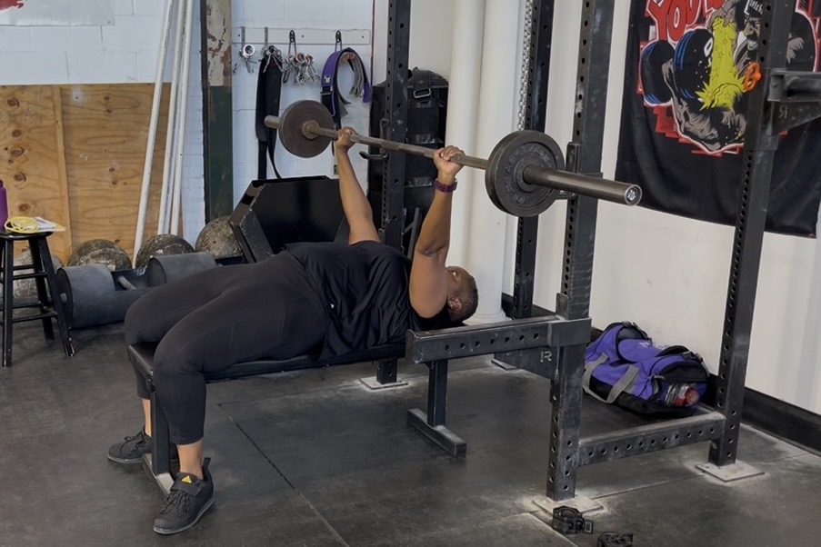 Tami Zanele bench pressing a barbell, wearing all black, focused and present during her Friday training session.