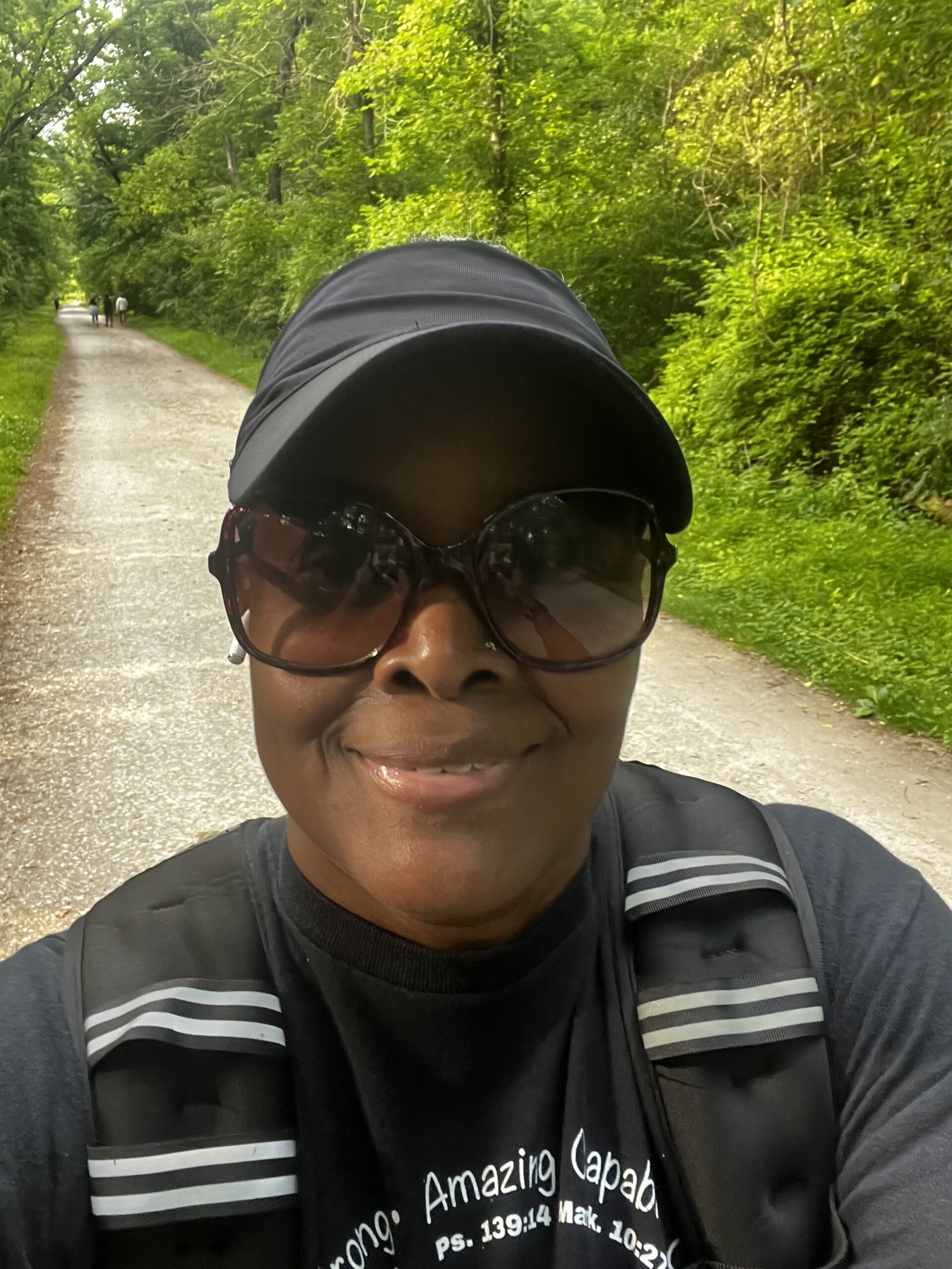 Tami Zanele smiling on a lush green nature trail, wearing a black cap, sunglasses, and a shirt reading "Strong, Amazing, Capable" with Psalm 139:14