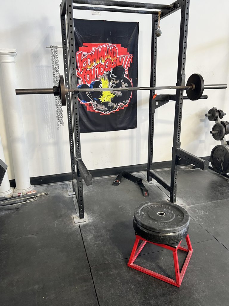 Barbell loaded to 100 pounds on a squat rack, with two black bumper plates stacked on a red box in front.